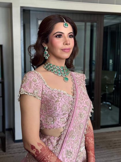 A portrait of the bride, her hair styled in soft, face-framing waves that complement her elegant pink lehenga and emerald jewelry.