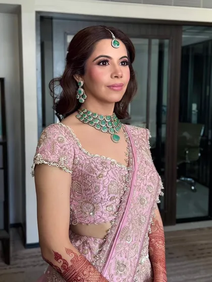 A portrait of the bride, her hair styled in soft, face-framing waves that complement her elegant pink lehenga and emerald jewelry.