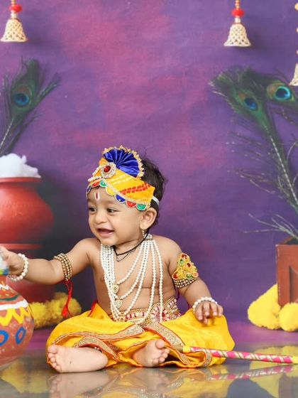 A playful moment as the baby Krishna reaches for a pot of "butter." The joyful expression and colorful backdrop make this a delightful and memorable photo.