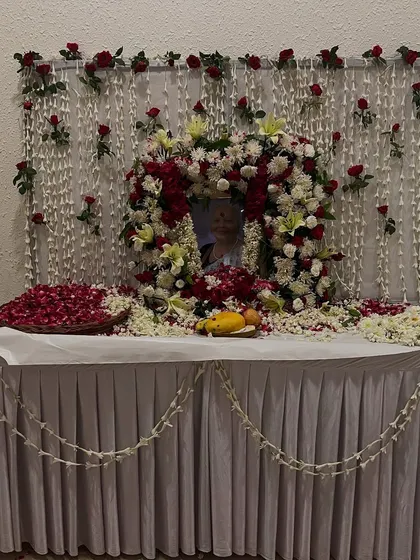 Another memorial setup with a beautiful floral wreath around the photograph. The table is decorated with a white cloth, scattered petals, and a garland of jasmine flowers.