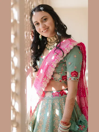 A radiant bridal portrait. The natural light from the window illuminates the bride's joyful expression and the beautiful details of her floral embroidered lehenga and traditional choker necklace.