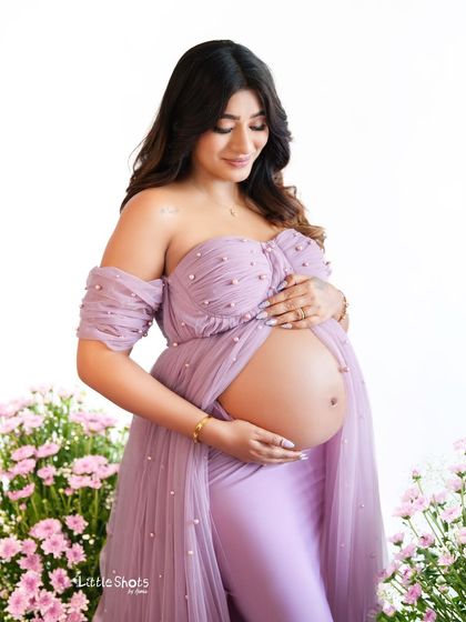 A beautiful shot focusing on the baby bump. The mother-to-be, in a lavender two-piece gown, cradles her belly, surrounded by a field of purple and pink flowers.