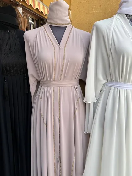 A trio of abayas in black, light pink, and white. These pieces show the range from classic and simple to styles with delicate gold chain detailing.