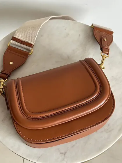 A close up of the Noble Leather Square Bag in a rich brown colour, laid out on a marble surface. You can see the quality of the smooth leather and the sturdy fabric shoulder strap that comes with it.