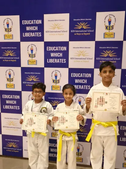 Three young karatekas from KES International School proudly display their new yellow belts and certificates. This is a significant first step in their martial arts training, marking their official entry into the world of karate.