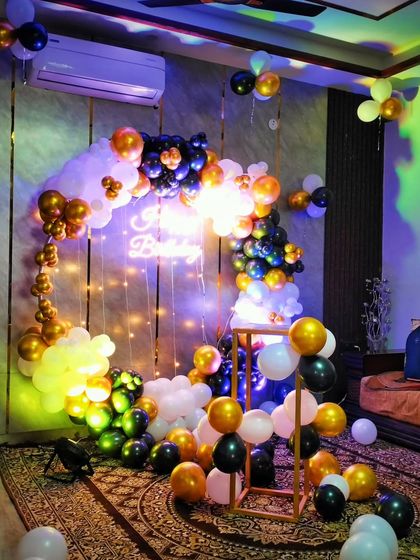 A dramatic birthday setup at home with a balloon arch in black, white, and gold. The multicolored lights and neon sign create a dynamic and club-like atmosphere.