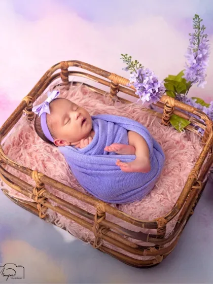 A perfect little bundle. This newborn is peacefully asleep in a rustic basket, surrounded by soft textures and delicate lavender flowers.