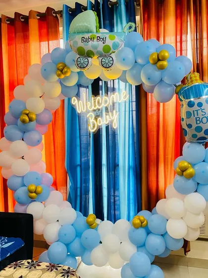 A classic "Welcome Baby" boy decoration. This setup features a circular balloon arch in blue and white, a neon sign, and foil balloons shaped like a baby carriage and a milk bottle.