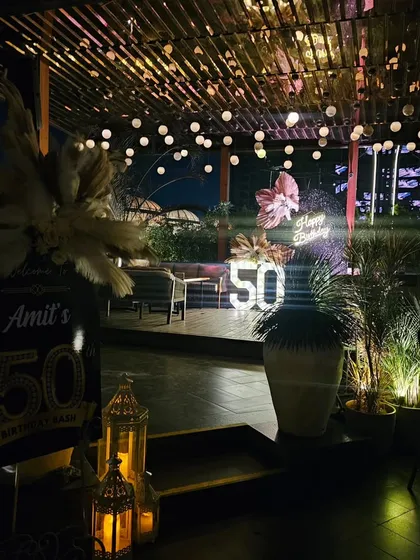 An elegant rooftop 50th birthday party at dusk. This image captures the beautiful ambiance we create with string lights, a stylish photo backdrop, and candle-lit lanterns for a memorable outdoor celebration.