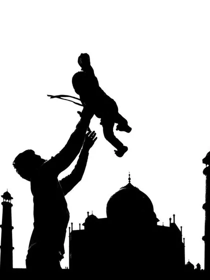A father lifts his child into the air, their silhouettes perfectly framed against the iconic outline of the Taj Mahal in a creative black and white composition.