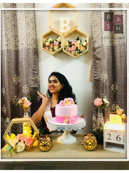 A sweet and floral-themed birthday moment. The birthday girl poses with her cake, surrounded by our decor featuring hexagonal shelves filled with flowers, a custom date calendar, and soft, warm lighting.