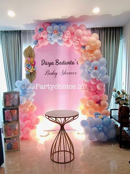 A lovely baby shower decoration for a home, using a pastel pink and blue balloon arch. The transparent "BABY" boxes filled with balloons add a popular and cute touch.