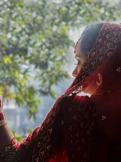 A moment of quiet anticipation. This photo shows the bride from behind, looking out a window. The natural light from outside silhouettes her form, highlighting the sheer red dupatta and the traditional chooda bangles on her arms.