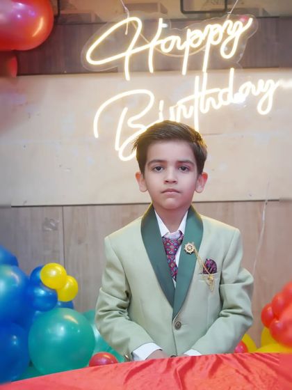 A young boy in a smart suit stands in front of a 'Happy Birthday' neon sign.