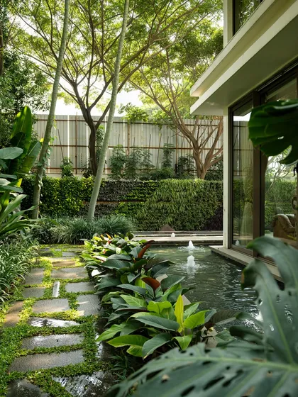 A lush, tropical garden path runs alongside a serene water body with fountains. The design uses a mix of stone pavers and ground cover, with the sound of water creating a peaceful journey through the space.