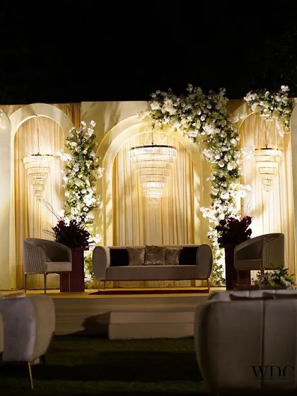 We design stages that are both grand and intimate. This regal reception stage features elegant arches, cascading white florals, and modern tiered chandeliers, creating a sophisticated backdrop for the couple's special evening.