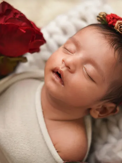 A close-up of a newborn girl's face, with a single red rose beside her. The contrast of the vibrant flower and her soft, sleeping features creates a striking and beautiful portrait.