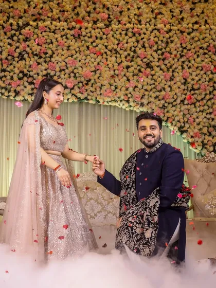 The moment after the proposal. The groom-to-be holds his fiancée's hand, both smiling, on a stage filled with fog and flowers.
