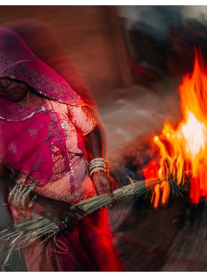 A woman in a pink saree stands near the Holika Dahan fire. The motion blur effect blends her with the flames, creating an abstract and powerful image of faith and ritual.