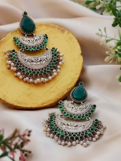 The natural texture of a wooden slice and fresh greenery provides an earthy backdrop for these oxidized silver and green stone earrings. This styling emphasizes the handcrafted, traditional feel of the jewelry.