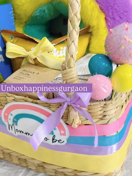 This image shows the variety of treats and decorative elements in the rainbow hamper, including mini balloons and a "Mom-to-be" sticker, all tied together with a lavender ribbon.