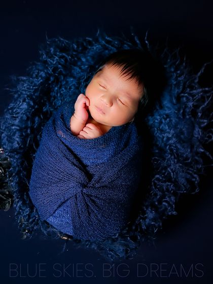 A newborn swaddled snugly in a rich blue wrap, nestled in a matching fur-lined bowl. The deep color creates a dramatic and beautiful contrast with the baby's soft skin.