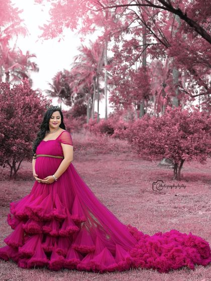 A regal portrait of a mother-to-be in a wine-colored ruffled gown and crown, set in a beautiful pink-themed garden.