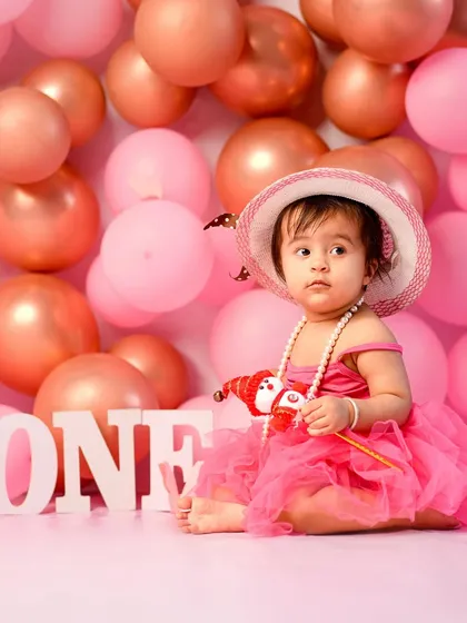 Pretty in pink for her first birthday. This portrait captures a sweet, thoughtful moment before the cake smash begins, showcasing her adorable outfit and the festive balloon backdrop.