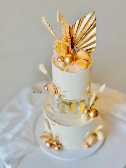 A sophisticated two-tier cake for a 60th birthday celebration in an ivory and gold theme. This design features a clear separator tier, macarons, and dried palm leaves for a modern, celebratory feel.