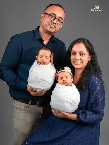 A classic posed portrait of a family with their newborn twins. The parents smile at the camera while securely holding their sleeping babies, creating a perfect first family photograph.