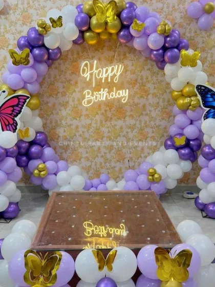 A butterfly-themed balloon ring in purple, white, and gold. The decoration features large butterfly cutouts on the balloon columns and smaller butterflies scattered throughout the ring for a whimsical feel.