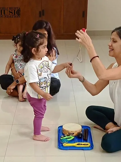 A teacher introduces a toddler to the triangle, a simple percussion instrument. Early exposure to different instruments helps develop a child's listening skills and curiosity.