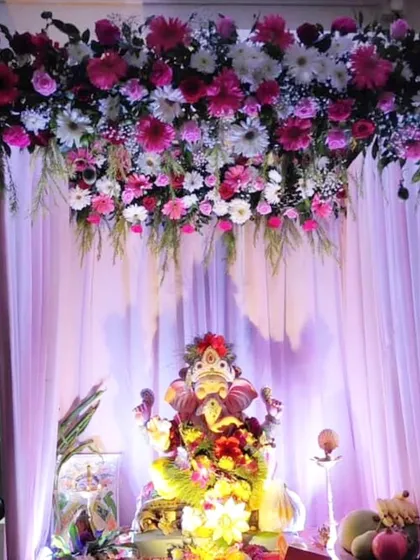 A beautiful hanging floral arrangement of pink and white gerberas and daisies creates a canopy effect over the Ganpati idol. The soft purple lighting enhances the festive mood.