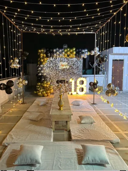 A stylish rooftop 18th birthday party. The setup includes a sequin wall with a black and gold balloon garland, low seating with cushions, and a canopy of fairy lights.