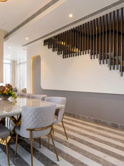 This shot showcases the custom staircase railing that acts as a decorative screen for the dining area. The vertical lines of the railing contrast with the horizontal stripes of the marble floor, creating a dynamic visual.