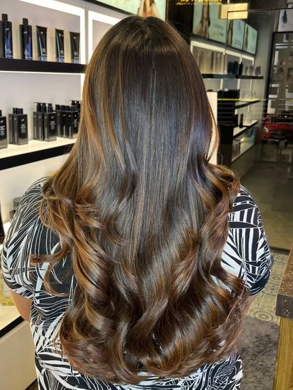 This image shows a deep brunette base with rich, warm highlights. The curls at the end add a touch of glamour, and the salon's product display in the background shows the premium brands I trust.