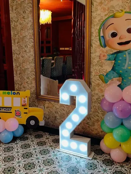 I use light-up marquee numbers to highlight the birthday age, seen here with a Cocomelon character balloon pillar and a themed school bus prop.