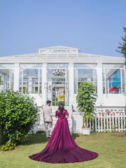 A fairytale moment as the couple approaches a stunning glasshouse conservatory. The bride's dramatic, long-trained gown creates a breathtaking silhouette, promising a story of modern elegance.
