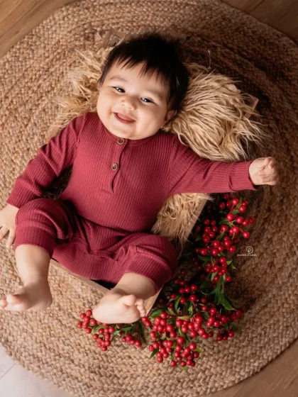 A happy baby in a maroon romper lies in a wooden crate on a jute rug. The simple, rustic props and pops of red berries create a warm and earthy portrait.