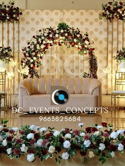 A beautiful reception stage combining a patterned backdrop with rich burgundy and white floral arrangements. The floor-level flowers create a seamless, garden-like feel, drawing all eyes to the main stage.