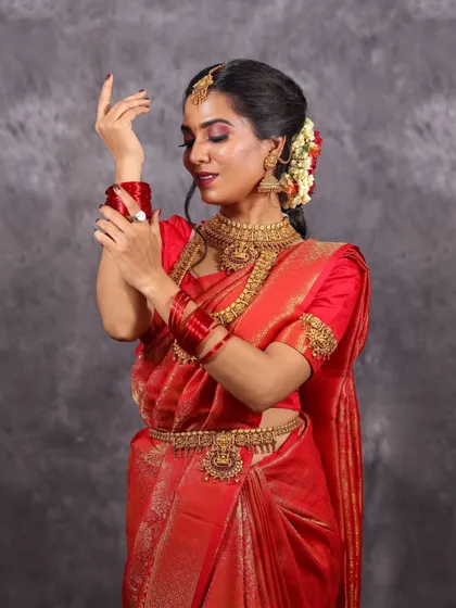 A classic bridal portrait pose, showing the model adjusting her bangles. This shot highlights the complete set of accessories, including the red bangles that match the saree.