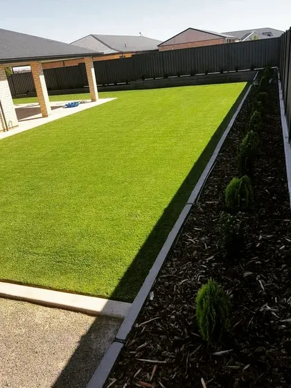 A simple and practical backyard with a neat artificial lawn and a border of mulch with small shrubs. This is a great example of a low-maintenance landscape solution.