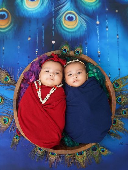 A vibrant and culturally rich portrait of twins. Dressed as Radha and Krishna, they look directly at the camera from their nest of peacock feathers.