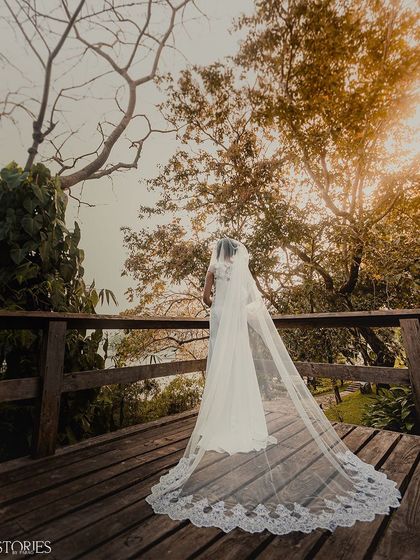 A bride in a flowing white gown with a long, elegant veil stands on a wooden deck overlooking the trees. This shot captures the grandeur and romance of a Christian wedding ceremony.