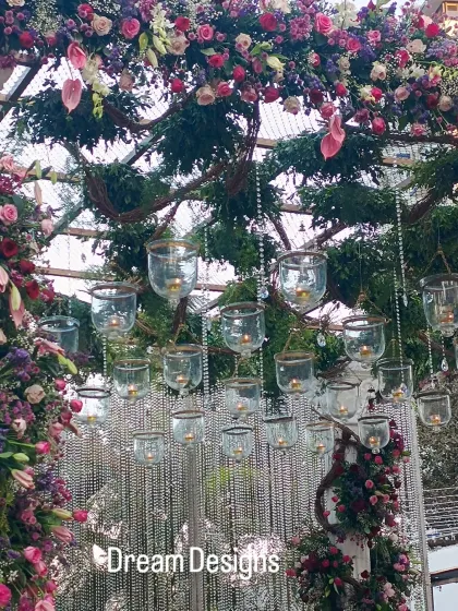 A close-up of a floral arch, showing the intricate details of the arrangement. Hanging glass tea light holders and crystal strings are nestled among the flowers, adding a delicate sparkle.
