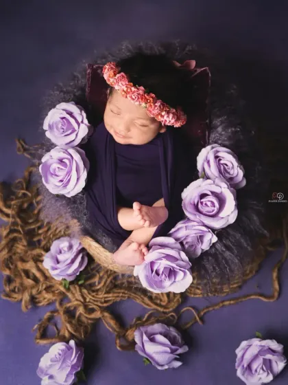 A sleeping newborn is nestled in a basket, surrounded by beautiful purple roses against a rich purple backdrop. The floral crown adds a final touch to this artistic composition.