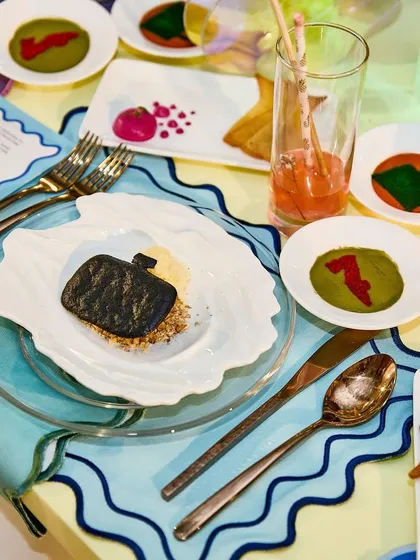 A dish featuring a charcoal cookie shaped like a handbag, part of a creative menu for a luxury fashion event. This plate shows our commitment to playful, thematic food design that directly references the world of couture.