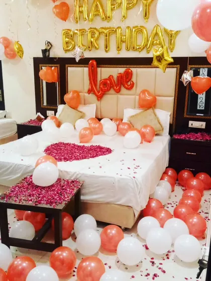 A bright and cheerful birthday room setup with rose gold, white, and gold balloons. The decor features 'Happy Birthday' and 'Love' foil balloons, and a rose petal heart on the bed.
