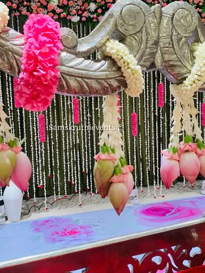 A close-up of a silver cradle decorated with hanging lotus buds and pink floral garlands. The backdrop of hanging jasmine strings adds a delicate and fragrant touch to the setup.