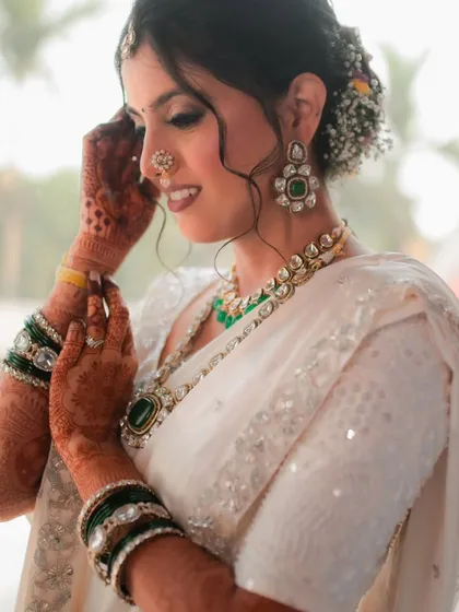 The bride adjusts her earring, giving a glimpse of her intricate floral hair bun and the heavy kundan and emerald jewelry that beautifully complements her ivory saree.